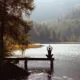 Person meditating on a dock by a serene lake surrounded by trees.