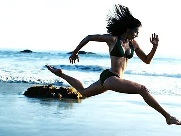 Woman in bikini jumping energetically over water near rocks.