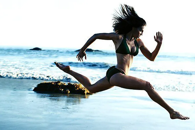 Woman in a bikini jumping over water near rocks.